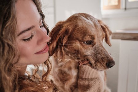 Téléchargez les photos : Heureux jeune propriétaire femelle avec les cheveux bouclés souriant et regardant à chien brun mignon à la maison - en image libre de droit