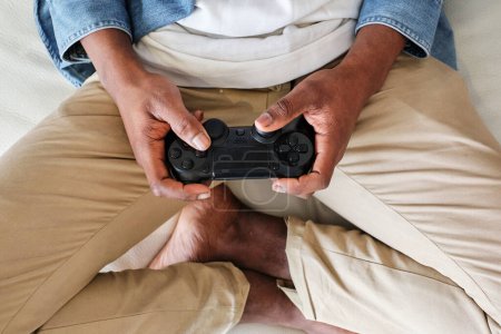 Photo for From above crop anonymous African American male gamer wearing casual outfit playing video game with gamepad while sitting on soft couch with legs crossed - Royalty Free Image