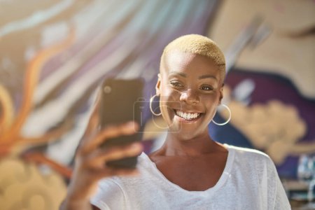 Foto de Sonriente joven afroamericana con el pelo corto teñido y pendientes en camiseta blanca tomando selfie en el teléfono inteligente - Imagen libre de derechos