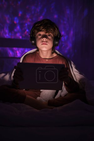 Téléchargez les photos : Garçon mignon dans les écouteurs regardant loin tout en naviguant tablette moderne sur le lit dans la chambre à coucher avec une lumière tamisée la nuit dans l'appartement - en image libre de droit