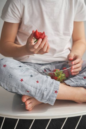 Photo for Unrecognizable barefoot boy in sleepwear sitting on white table and eating sweet berries from bowl at home on weekend day - Royalty Free Image