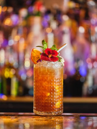 Téléchargez les photos : Cocktail alcoolisé décoré de fleurs comestibles et d'orange séchée servi en verre avec glaçons au bar sur fond flou - en image libre de droit