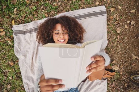 Photo for Top view of black woman with Afro hairstyle reading interesting story and smiling while resting on blanket on grass in nature - Royalty Free Image