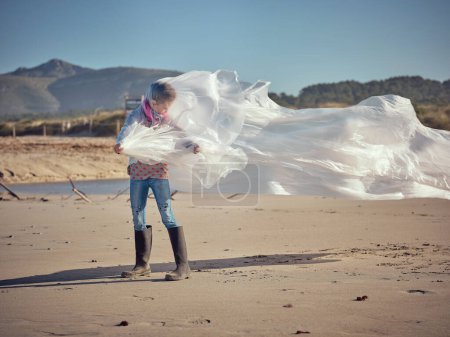 Foto de Niño irreconocible parado solo en la playa de arena en un día soleado y ventoso y mirando hacia otro lado mientras está envuelto en una sábana blanca - Imagen libre de derechos