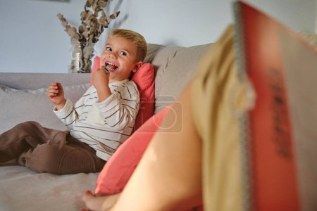 Photo for Adorable boy resting on sofa near unrecognizable woman and playing with whistle in living room at home on weekend day - Royalty Free Image