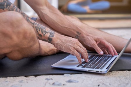 Téléchargez les photos : Homme tatoué anonyme assis sur le tapis et tapant sur le clavier de l'ordinateur portable pendant l'entraînement dans la salle de gym - en image libre de droit