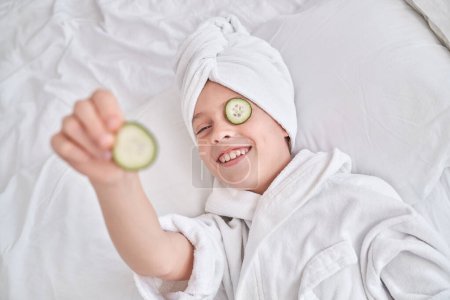 Téléchargez les photos : D'en haut de l'enfant en turban blanc et peignoir souriant et regardant la caméra tout en se relaxant sur un lit doux et en démontrant une tranche de concombre lors des procédures de spa à la maison - en image libre de droit
