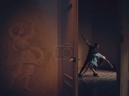 Téléchargez les photos : Vue latérale d'un homme anonyme et d'une tendre ballerine dansant dans une pièce vide derrière une porte ouverte dans une lumière tamisée - en image libre de droit