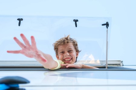 Foto de Desde abajo niño alegre sonriendo felizmente y alcanzando la cámara mientras se asoma por la ventana de RV en el soleado día de verano - Imagen libre de derechos