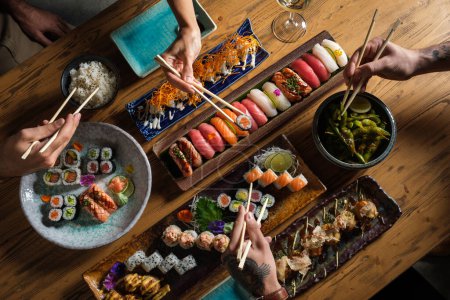 Photo for From above of friends eating tasty various sushi with chopsticks while sitting at table with rice and green peas - Royalty Free Image