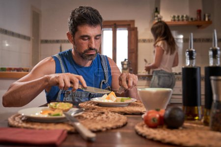Téléchargez les photos : Focalisé barbu homme manger appétissant oeufs brouillés tandis que la femme cuisine en arrière-plan à la maison dans la cuisine - en image libre de droit