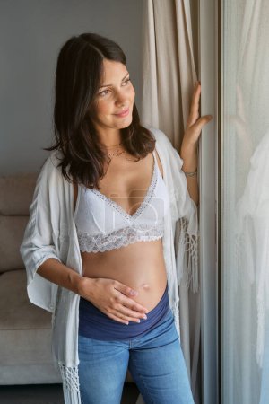 Photo for Black haired satisfied lady in white brassiere looking out window in living room while touching pregnant tummy - Royalty Free Image