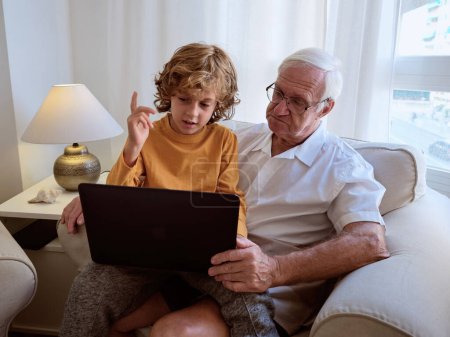 Photo for Senior man with cute grandson in casual wear browsing netbook together while sitting in comfortable armchair near window at home - Royalty Free Image