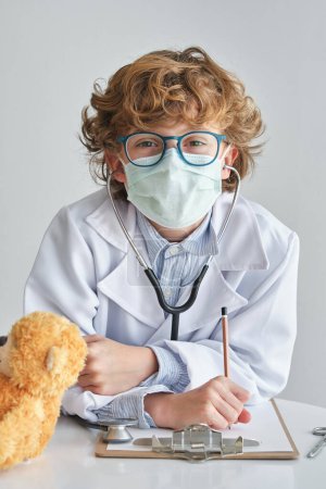 Photo for Confident child in medical uniform and eyeglasses looking at camera. Leaning on table with soft bear and clipboard on white background - Royalty Free Image