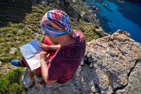 Téléchargez les photos : D'en haut du voyageur méconnaissable en bandana vérifier la carte tout en étant assis sur une falaise de montagne rocheuse pendant le voyage d'été - en image libre de droit