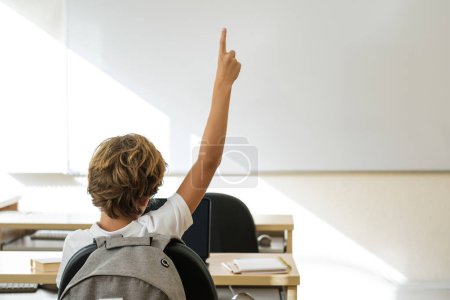 Foto de Vista posterior del niño irreconocible con el brazo levantado sentado en la mesa en el aula durante la lección - Imagen libre de derechos