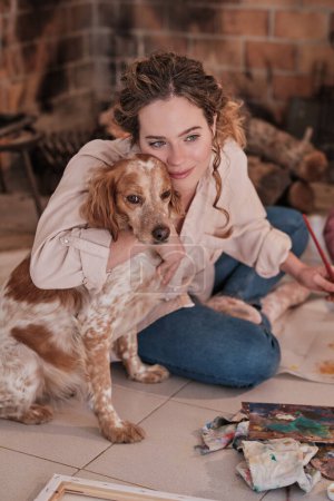 Téléchargez les photos : Artiste souriante assise près de la cheminée et caressant chien espagnol tout en peignant dans un studio moderne à la maison - en image libre de droit