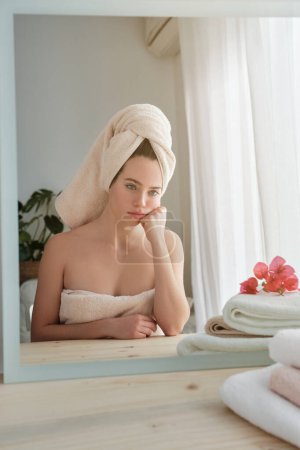Foto de Mujer con el pelo envuelto en toalla blanca sentada en la mesa de madera y soñando despierto mientras mira a la reflexión después de hacer la rutina de belleza matutina en casa - Imagen libre de derechos