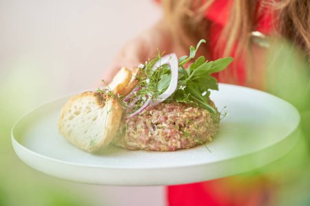 Photo for High angle of crop anonymous female holding delicious steak tartar with green herbs and red onion served on white plate - Royalty Free Image