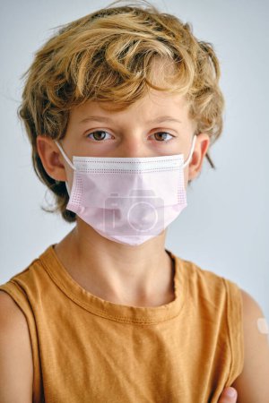 Foto de Niño serio con cabello castaño en máscara médica mirando a la cámara durante la pandemia de COVID 19 - Imagen libre de derechos