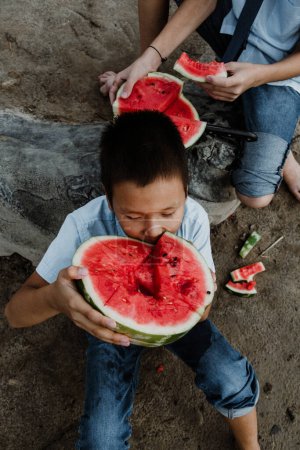 Photo for From above of crop male children in school uniform sitting on sandy ground and eating delicious watermelon in daylight - Royalty Free Image