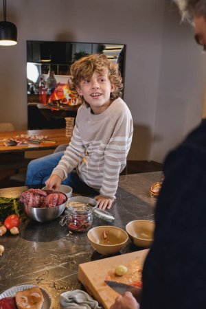 Téléchargez les photos : Bouclé garçon souriant en pull assis sur un large comptoir de cuisine plein d'ustensiles de cuisine contre père - en image libre de droit