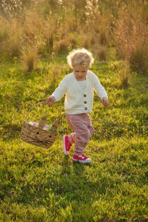 Foto de Cuerpo completo linda niña con el pelo rizado usando traje elegante paseando por prado herboso en la naturaleza de verano y mirando hacia abajo - Imagen libre de derechos