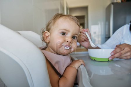 Photo for Curious baby girl in bib sitting on high chair and looking at camera with interest while eating with help of crop woman in kitchen at home - Royalty Free Image
