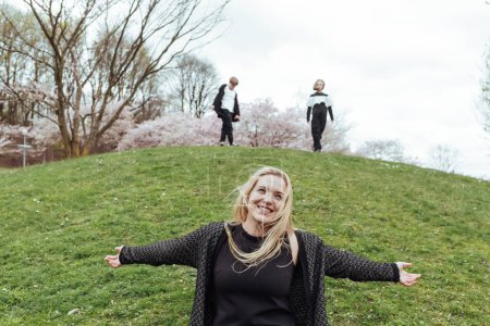 Foto de Sonriente madre rubia en la colina verde cerca de los niños y árboles florecientes en el parque - Imagen libre de derechos
