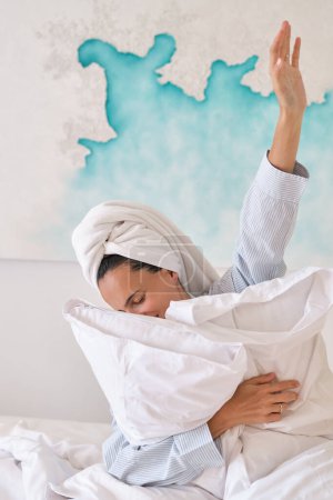 Foto de Feliz joven hembra con toalla envuelta alrededor de la cabeza abrazando almohadas blancas sentadas en la cómoda cama en casa - Imagen libre de derechos