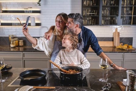 Photo for Cheerful diverse parents in aprons cooking together with preteen son in kitchen and taking self portrait on smartphone - Royalty Free Image
