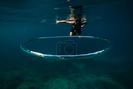 Foto de Cuerpo completo de joven buzo sin camisa en pantalones cortos aletas y máscara de snorkel flotando bajo el agua del mar cerca de la burbuja toroidal cerca de los arrecifes de coral - Imagen libre de derechos