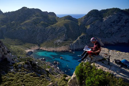 Téléchargez les photos : Vue latérale du voyageur masculin en vêtements décontractés assis sur une falaise rocheuse et lisant une carte en papier par temps ensoleillé - en image libre de droit