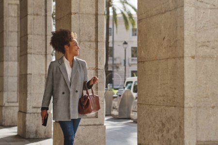 Téléchargez les photos : Femme afro-américaine positive en vêtements décontractés intelligents regardant loin sur la rue de la ville tout en se rendant au travail en plein jour - en image libre de droit