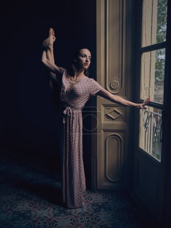 Foto de Mujer en vestido elegante y punta de pie cerca de la puerta y mirando hacia otro lado mientras sostiene la pierna hacia arriba en la luz tenue - Imagen libre de derechos
