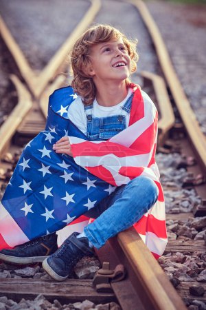 Téléchargez les photos : Enfant satisfait enveloppé dans le drapeau des États-Unis et levant les yeux assis sur le chemin de fer pendant la célébration de la fête de l'indépendance - en image libre de droit