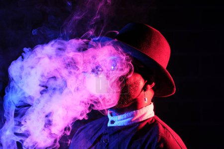 Téléchargez les photos : Vue latérale du jeune homme noir en tenue élégante et chapeau debout dans une pièce sombre avec de la fumée - en image libre de droit