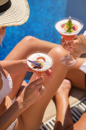 Photo for High angle of anonymous woman in sunhat and swimwear enjoying delicious cocktail while sitting at pool during summer day - Royalty Free Image