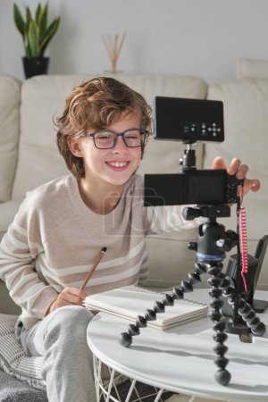 Foto de Niño sonriente con el pelo rizado configurar la cámara para grabar vídeo para el blog en línea en la sala de estar moderna en casa - Imagen libre de derechos