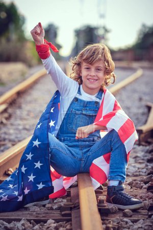 Foto de Cuerpo completo de niño positivo envuelto en colorida bandera de EE.UU. mirando a la cámara con el brazo levantado mientras está sentado en el ferrocarril de metal - Imagen libre de derechos