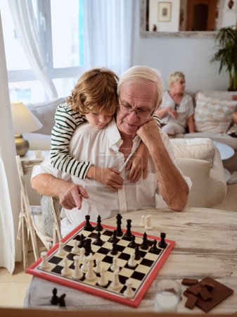 Téléchargez les photos : Preteen garçon étreignant homme âgé et apprenant à jouer aux échecs à table tandis que grand-mère assis sur le canapé dans le salon confortable à la maison - en image libre de droit