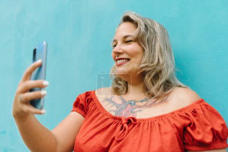 Photo for Portrait of young plus size female in casual clothes taking selfie with smartphone while standing on blue wall background outdoors - Royalty Free Image
