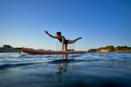 Téléchargez les photos : Corps complet de la femme concentrée en maillot de bain debout sur le tableau SUP en équilibre pose tout en pratiquant le yoga dans l'ondulation de la mer - en image libre de droit