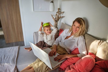 Photo for Side view of positive woman typing on laptop and laughing at cheerful boy putting green plastic bowl on head while sitting on couch in living room at home - Royalty Free Image