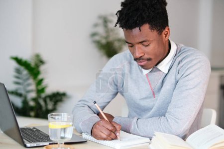 Téléchargez les photos : Afro-Américain prenant des notes dans le copybook entre netbook et livre tout en étudiant à table dans la maison - en image libre de droit
