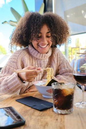 Photo for Content young African American female with curly hair in sweater sitting at wooden table while eating ramen with chopsticks - Royalty Free Image