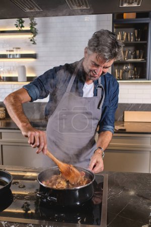Téléchargez les photos : Chef masculin souriant debout à la cuisinière et en remuant les pâtes dans la cuisine moderne à la maison - en image libre de droit