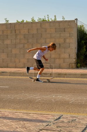 Téléchargez les photos : Vue latérale du patineur préadolescent énergique chevauchant le skateboard sur la route dans la soirée ensoleillée et divertissant pendant le week-end d'été - en image libre de droit