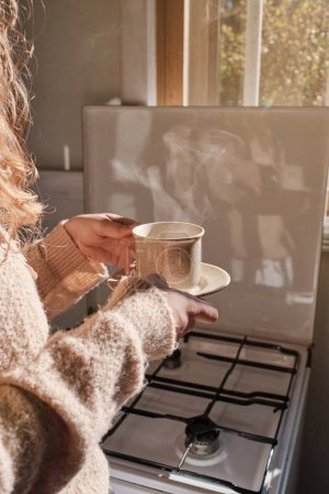 Foto de Cultivo irreconocible mujer anónima en suéter caliente con taza de bebida caliente en la cocina en casa - Imagen libre de derechos