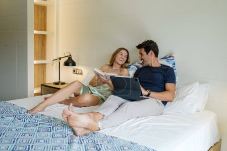 Photo for Smiling girlfriend reading book on bed near cheerful boyfriend browsing tablet while spending weekend together in bedroom - Royalty Free Image